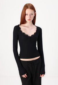 Black ribbed long-sleeve top with a scalloped lace neckline and button detail, featuring fitted sleeves with thumbholes.