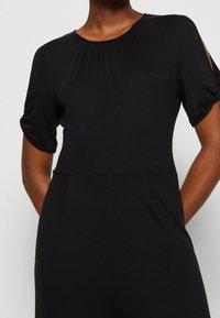 Černý overal s krátkými, ohrnutými rukávy. Má skládací výstřih a hladký povrch, s viditelnými bočními otvory na rukávech.