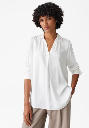 Chemise blanche à manches longues avec un col en V et des poignets retroussés ; tissu lisse et léger ; coupes décontractées et ourlet arrondi. Associée à un pantalon beige.