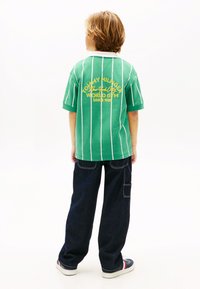 Πράσινο κοντομάνικο πουκάμισο με άσπρες ρίγες και κίτρινο κείμενο στην πλάτη. Σκούρο μπλε φαρδιά τζιν παντελόνια ολοκληρώνουν την εμφάνιση.