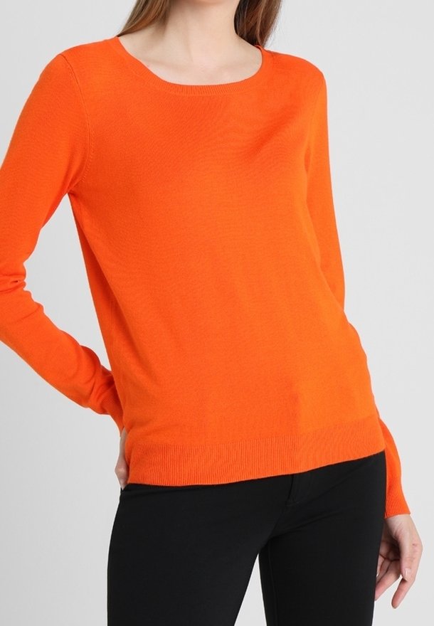 Femme portant un pull orange vif à manches longues et un pantalon noir, debout avec une main dans le dos sur un fond uni.
