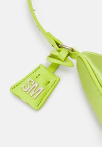 Accessoire en cuir vert avec une étiquette en métal argenté brillant portant le marquage "SM" en relief. Le design présente une texture lisse et une forme élégante.