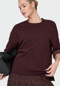 Bordowy sweter z dzianiny o okrągłym dekolcie i luźnym kroju, zestawiony z wzorzystą spódnicą, trzymaną w czarnej skórzanej torbie.