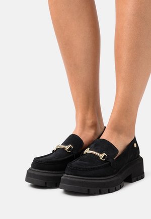 Mocassins en suède noir avec une semelle en caoutchouc noir épaisse, présentant un accent en barrette dorée sur l'empeigne et un design à bout rond.