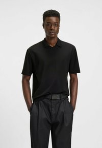 Schwarzes Polohemd mit V-Ausschnitt und kurzen Ärmeln, kombiniert mit hochgeschnittenen schwarzen Hosen und einem schwarzen Gürtel mit Logodetails.