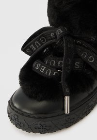 Black ankle boot with faux fur trim and thick black rubber sole, featuring logo-printed black laces and metallic eyelet accents.