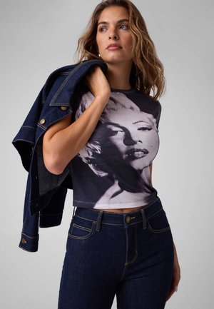 Vrouw draagt donkere jeans en een cropped t-shirt met een zwart-witte gezichtprint, houdt een spijkerjasje over haar schouder.