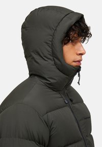 Mørkegrøn dunjakke med hætte, udstyret med lynlås foran, ribbede sømme og en glat tekstur. Detaljer inkluderer justerbare snore ved halsen.