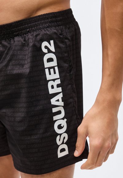 Μαύρο σορτς με ελαστική μέση, διαθέτει κατακόρυφο λευκό λογότυπο "DSQUARED2" στο πλάι, φορεμένο από άτομο με εμφανές χέρι και κορμό.