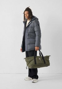 Giovane con giacca imbottita grigia e pantaloni neri, che tiene una grande borsa a tracolla verde oliva con manici neri, in piedi contro un muro bianco.