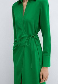 Grünes Wickelkleid aus glattem Stoff. Verfügt über einen Kragen, lange Ärmel, einen Taillengürtel und geraffte Details auf einer Seite. Schlitz im Rock.