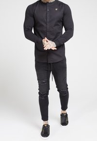 Hombre con tatuajes visibles en las manos, vistiendo una camisa negra de botones, unos jeans skinny negros rasgados y zapatillas negras con acentos dorados.