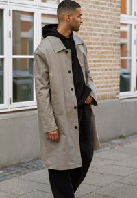 Lys grå trenchcoat med bred krave, knaplukning foran og lange ærmer, båret over en sort hættetrøje og mørke bukser. Glat stoffinish.