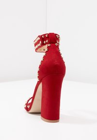 Sándala de tacón alto de ante rojo con un tacón grueso, correa en el tobillo y tachuelas doradas a lo largo de los bordes, mostrando una superficie texturizada.