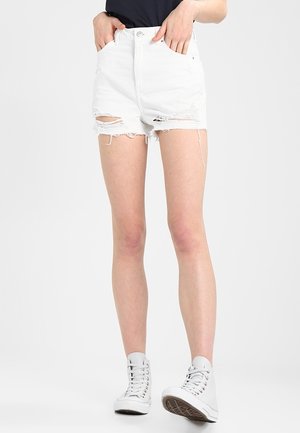 Personne portant un short en jean blanc déchiré, un haut noir et des baskets montantes blanches, debout les mains dans les poches.