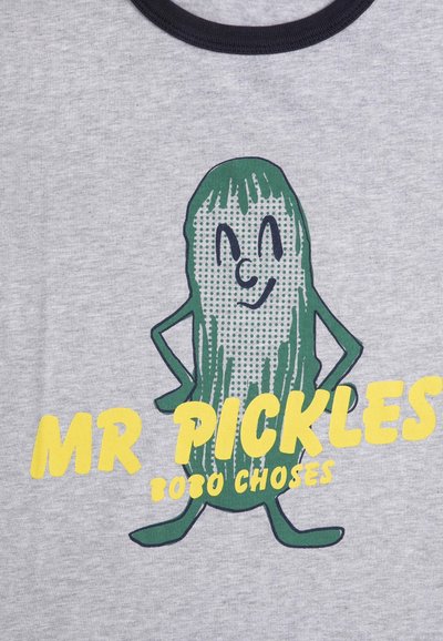 Cartoon grüner Gurken mit lächelndem Gesicht und Händen in die Hüften gestützt, gelber Text "MR PICKLES" und kleinerer Text "TOTO CHOSES" auf grauem Stoff.