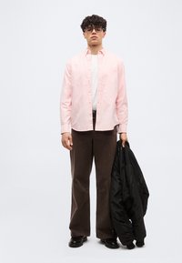 Jeune homme aux cheveux bouclés et lunettes, portant une chemise rose clair, un pantalon marron, des chaussures noires, tenant une veste noire dans une main.