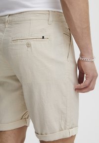 Beige vászonshort, felgombolt szegéllyel, hátul gombos zsebbel és finom logóhímzéssel. Sima textúra és lazább kialakítás.