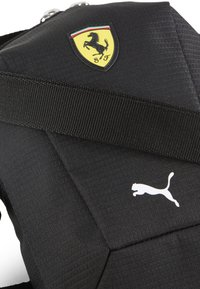 Sort rygsæk med tekstureret stof, prydet med et gult Ferrari-logo og et hvidt Puma-logo. Indeholder en holdbar, justerbar rem.