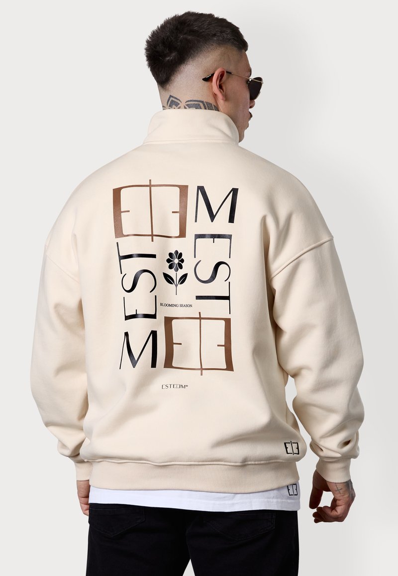 Sweatshirt surdimensionné couleur crème avec un col montant, présentant une grande impression graphique en noir et marron au dos, incluant du texte et un motif de fleur.