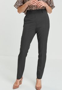 Femme portant un pantalon noir ajusté, un chemisier à motif léopard et des chaussures à talons hauts marron, se tenant devant un fond uni.