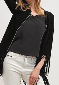Black suede jacket with fringe details, silver zipper, worn over a black top. White jeans visible, showcasing a fitted silhouette.