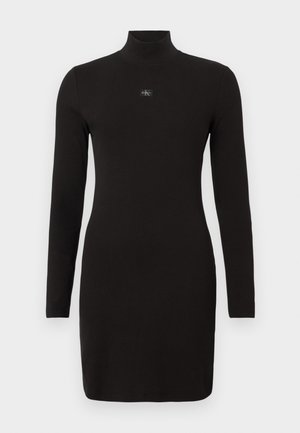 Vestido preto de malha canelada com mangas compridas e gola alta, apresentando um pequeno emblema com o logotipo na parte frontal. O tecido aparenta ser macio e suave.