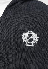 Μαύρο πλεκτό φούτερ με υφασμάτινη υφή, διακοσμημένο με λευκό κεντητό λογότυπο στο στήθος. Το σχέδιο περιλαμβάνει κουκούλα και ριμπ άκρα.