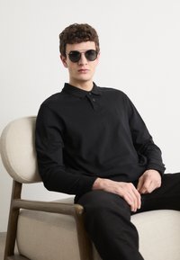 Juoda ilgomis rankovėmis polo marškiniai su trimis mygtukais. Modelis nešioja tamsius akinius nuo saulės, sėdi ant šviesiai nusidažytos, audiniu aptrauktos kėdės.