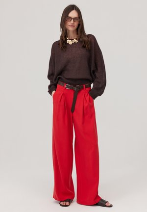 Kobieta w ciemnobrązowym, luźnym swetrze, szerokich czerwonych spodniach z czarnym paskiem, czarnych sandałach, złotym naszyjniku i okularach przeciwsłonecznych, stoi z rękami w kieszeniach.