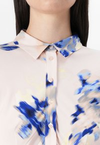 Närbild av en kvinnas krage med en blå och beige mönstrad blus. Kragen är knäppt och har konstnärliga, abstrakta blommönster.