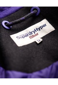 Sort stof med lilla accenter, der har et mærke med teksten "SuperdryHyper Biker," størrelse S, med en flettrukket lilla snor.