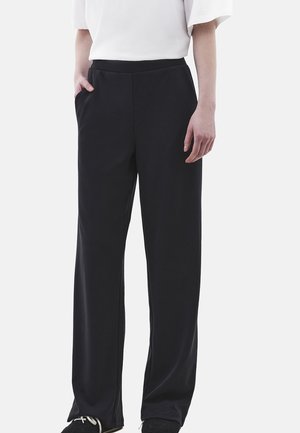 Osoba nosząca luźne czarne spodnie z bocznymi kieszeniami, biały top i czarne trampki, stojąca na tle jednolitego jasnego tła.