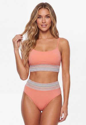 Kvinde med langt bølget blondt hår iført en koralfarvet todelt badedragt med brede flerfarvede zigzag-mønstrede bånd ved taljen og under brystet.