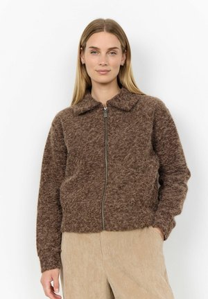 Frau mit glattem blondem Haar, die eine braune flauschige Reißverschlussjacke und beigen Cordhose trägt, steht mit einer Hand in der Tasche vor weißem Hintergrund.