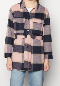 Kobieta w różowej, granatowej i beżowej kraciastej kurtce z paskiem, z dużymi przednimi kieszeniami, nosi białą koszulę i niebieskie dżinsy.