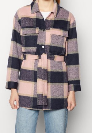 Vrouw in een roze, marineblauwe en beige geruite jas met riem en grote voorzakken over een witte blouse en blauwe jeans.