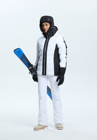 Skikleding bestaande uit een wit en zwart gewatteerd jack, witte broek, zwarte handschoenen en beige skibotten, met blauwe ski's met "WEDZE" in de hand.