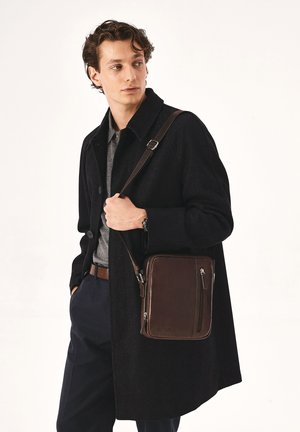 Hnedá kožená taška na rameno s obdĺžnikovým tvarom, ktorá má predné zipsové vrecko a nastaviteľný popruh, predvádzaná modelom v čiernom kabáte.