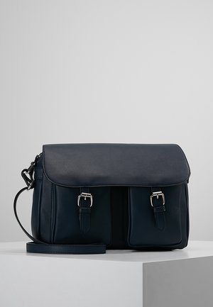 Sac bandoulière en cuir bleu foncé avec fermeture à rabat et deux poches avant à boucle, présenté sur une plateforme blanche sur fond gris.