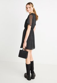 Μαύρο φόρεμα με κοντά διαφανή μανίκια, πουά σχέδιο, χαλαρή γραμμή και φουσκωτή φούστα· συνδυασμένο με μαύρες μπότες μέχρι το γόνατο και μια μικρή τσάντα.