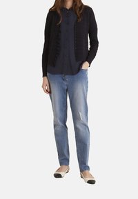 Námořnicky modrý pletený kardigan s volánky, zavírání na knoflík, kombinovaný s tmavým topem a světle modrými džíny. Obuv má krémový a černý design.