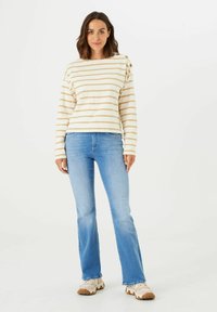 Gestreiftes Langarmshirt in Creme und Beige mit Knopfakzenten, kombiniert mit hellblauen Schlagjeans und cremefarbenen Turnschuhen.