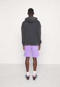 Sudadera gris con capucha, combinada con pantalones cortos lavanda. El calzado deportivo presenta un diseño multicolor. Fondo de color sólido.