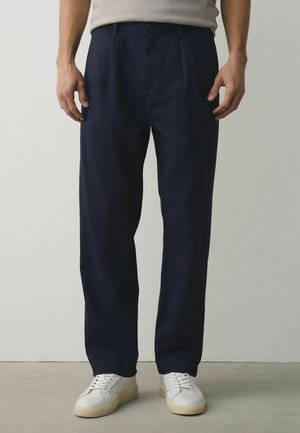 Man wearing navy blue straight-leg trousers, beige shirt tucked in, and white sneakers standing on gray floor against white wall.