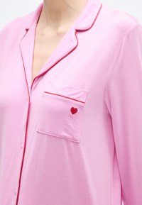 Bluza de pijama roz cu garnitură roșie, având un buzunar la piept și o broderie mică în formă de inimă roșie pe buzunar. Confecționată dintr-un material moale.