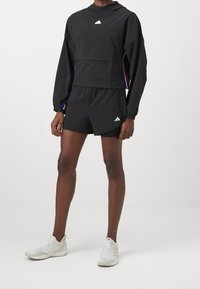 Conjunto deportivo negro de dos piezas que incluye una sudadera corta con capucha y bolsillo en el pecho, combinada con pantalones cortos elásticos y zapatillas blancas.