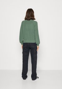 Maglione verde lavorato a maglia con texture a coste, maniche a raglan e orlo aderente, abbinato a pantaloni cargo scuri con tasche laterali.