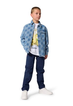 Veste en denim avec motif logo HUGO, t-shirt blanc avec imprimé graphique, pantalon marine et baskets blanches. Tenue décontractée et moderne.