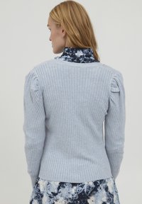 Pull en maille côtelée gris clair avec une silhouette ajustée, épaules bouffantes et col rond, superposé sur une chemise à motifs floraux sombres.